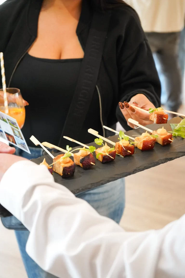 A woman holding a drink taking a portion from a decorative slate plate of individual pork belly skewers with garnish